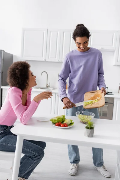 Cheerful man cooking salad near vegetables and girlfriend with coffee in kitchen — Foto stock