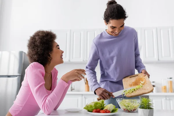 Jovem homem africano americano cozinhar salada perto de namorada sorridente com xícara de café na cozinha — Fotografia de Stock