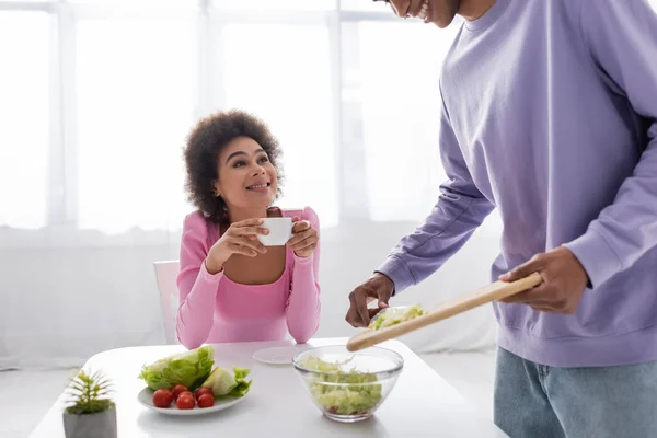 Giovane donna afro-americana in possesso di tazza vicino fidanzato cottura insalata in cucina — Foto stock