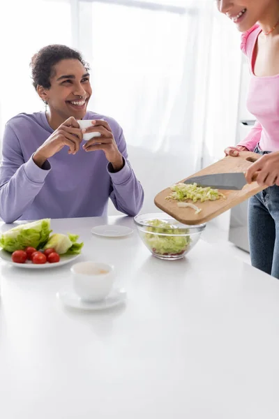 Smiling african american man holding cup while girlfriend cooking salad at home — Fotografia de Stock