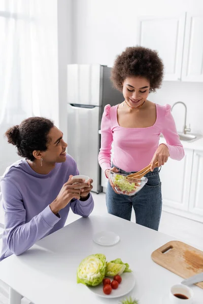 Young african american woman holding bowl with salad near boyfriend with coffee in kitchen — Stockfoto