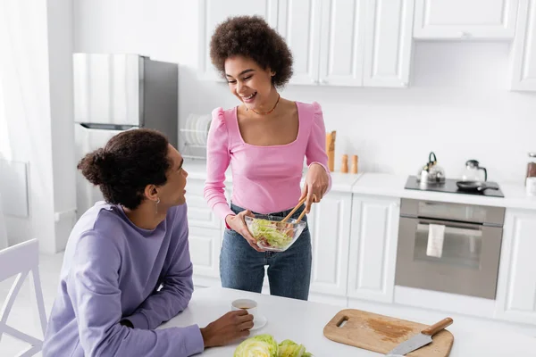 Positive african american woman holding bowl with salad near boyfriend with cup of coffee at home — Fotografia de Stock