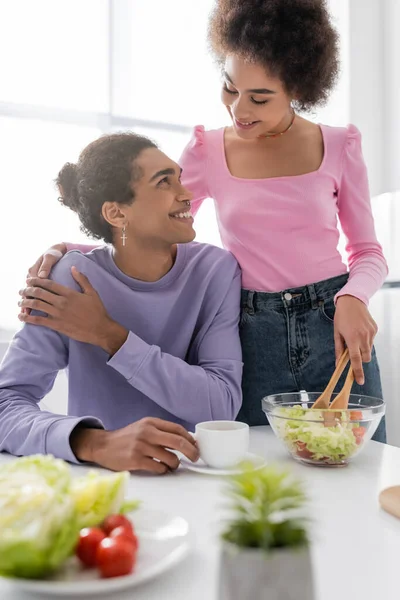 Smiling african american woman hugging boyfriend with cup while cooking salad in kitchen — Stockfoto