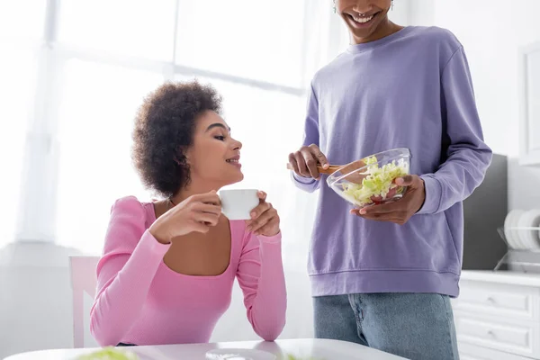 Joven mujer afroamericana sosteniendo taza cerca de novio con tazón de ensalada en casa - foto de stock