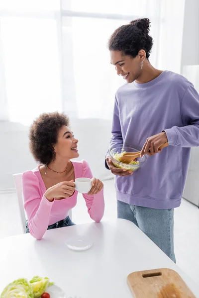 Allegro uomo africano americano che tiene ciotola con insalata vicino fidanzata con caffè a casa — Foto stock