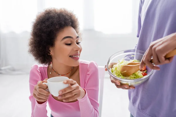 Positive Afroamerikanerin hält Tasse neben Freund, der zu Hause Salat mixt — Stockfoto