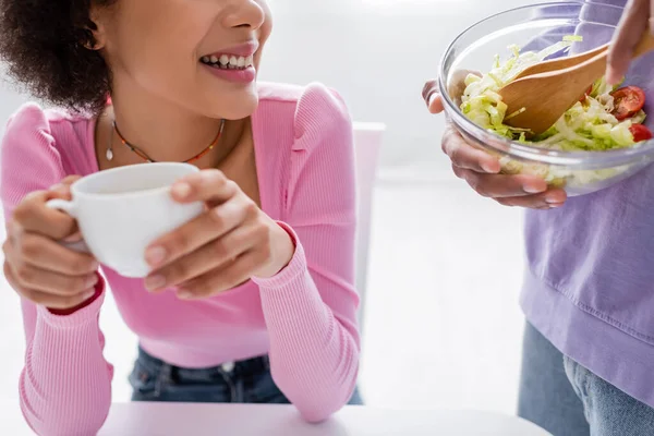 Vista recortada de la mujer afroamericana sosteniendo taza borrosa mientras su novio mezcla ensalada fresca en casa - foto de stock