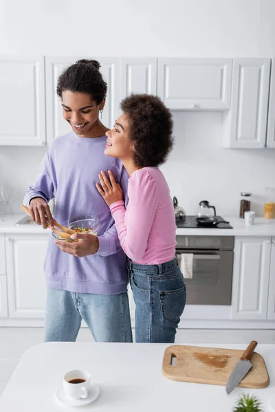 Alegre mujer afroamericana abrazando novio con tazón de ensalada cerca de la taza de café en casa - foto de stock