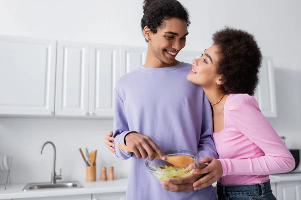 Sorridente uomo africano americano che tiene ciotola con insalata vicino fidanzata a casa — Foto stock