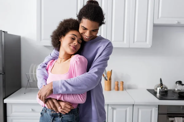 Alegre afroamericano hombre abrazando novia con los ojos cerrados en la cocina - foto de stock