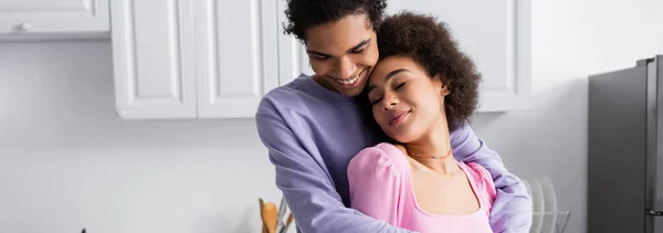 Positivo afroamericano hombre abrazando novia en casa, bandera - foto de stock