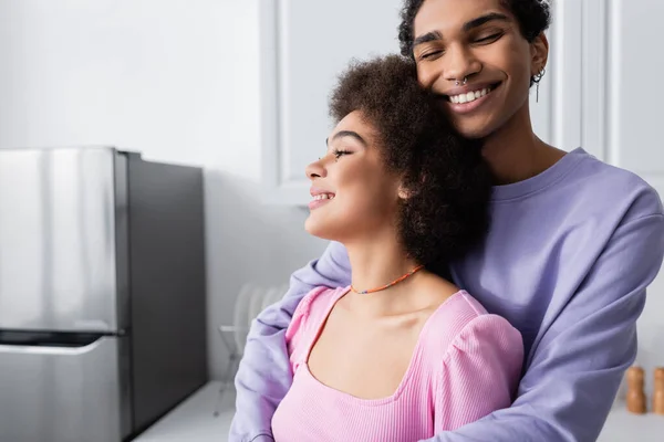 Heureux homme afro-américain avec les yeux fermés étreignant petite amie dans la cuisine — Photo de stock