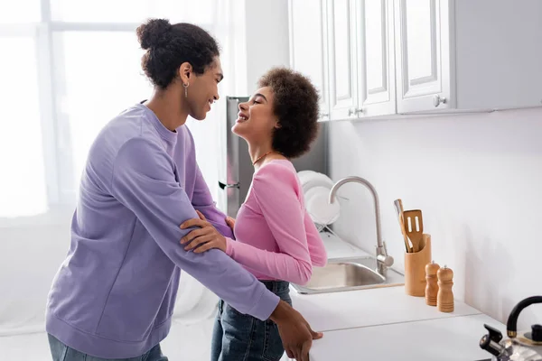 Vista lateral de la alegre pareja afroamericana mirándose en la cocina - foto de stock