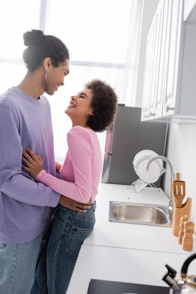 Vista lateral de la mujer afroamericana positiva abrazando novio cerca de encimera en la cocina - foto de stock
