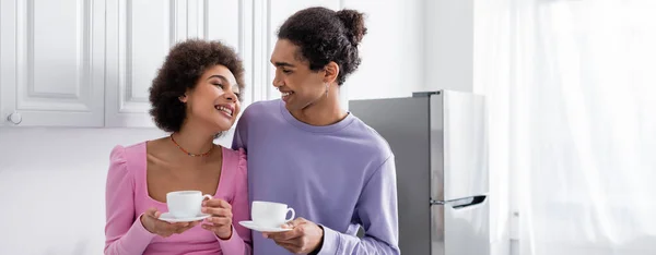 Sonriente mujer afroamericana sosteniendo taza de café cerca de novio en casa, pancarta - foto de stock