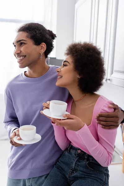 Positive african american man with cup hugging girlfriend in kitchen — Stockfoto