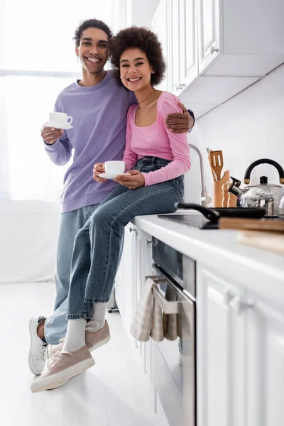 Joyeux couple afro-américain avec café étreignant et regardant la caméra dans la cuisine — Photo de stock