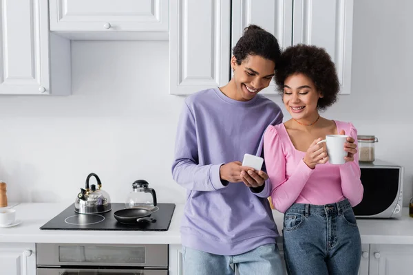 Positivo afroamericano hombre sosteniendo teléfono inteligente cerca de novia con taza en la cocina - foto de stock