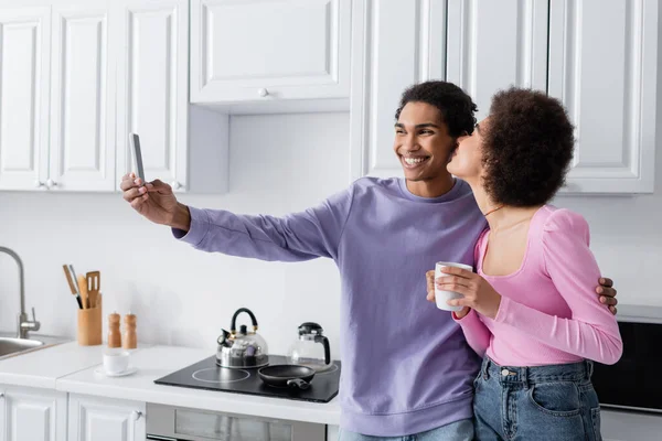 Smiling african american man taking selfie on smartphone near girlfriend with cup in kitchen — Stockfoto