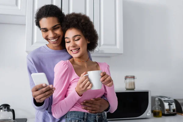 Heureux homme afro-américain en utilisant smartphone et étreinte petite amie avec tasse dans la cuisine — Photo de stock