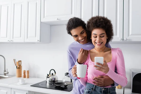 Mujer afroamericana positiva con taza usando teléfono inteligente cerca de novio en casa - foto de stock