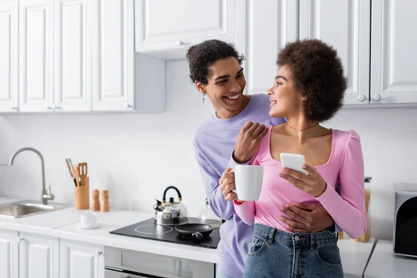 Молодая африканская американская пара со смартфоном и чашкой, смотрящая друг на друга на кухне — стоковое фото