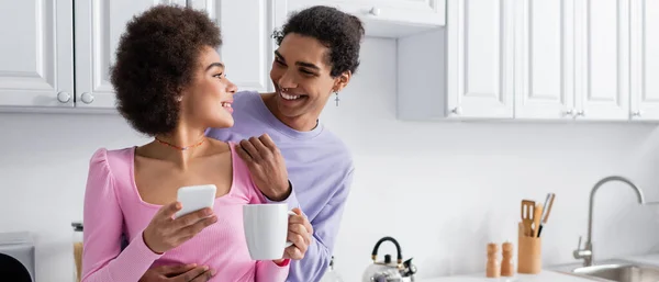 Hombre afroamericano abrazando novia con teléfono móvil y taza en la cocina, pancarta - foto de stock