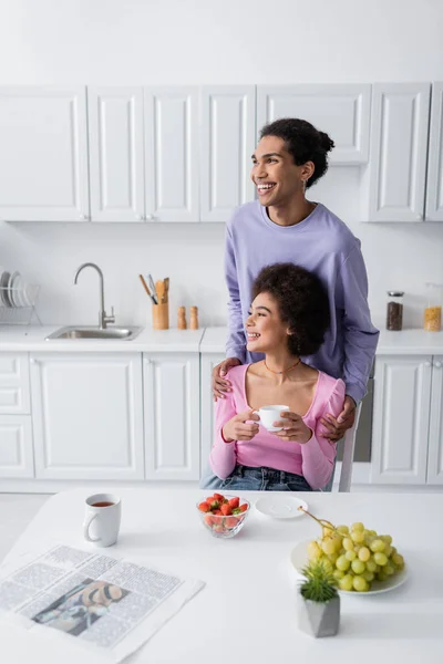Uomo afroamericano che abbraccia la fidanzata con tazza vicino alla frutta e giornale in cucina — Foto stock