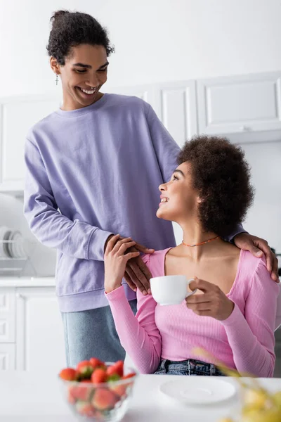 African american man hugging girlfriend with cup near blurred strawberries in kitchen — Stock Photo