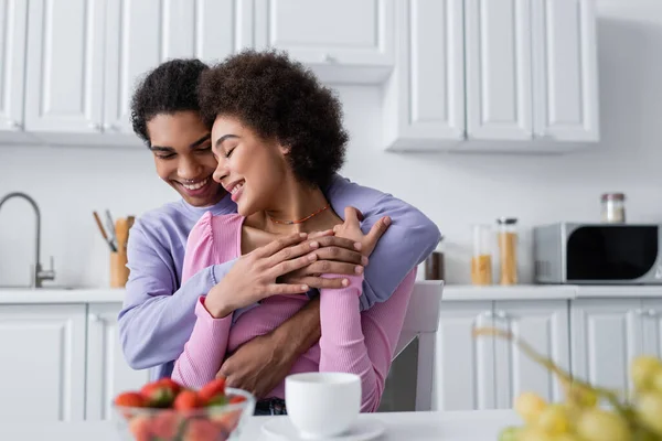 Glücklicher afrikanischer Amerikaner umarmt Freundin in der Nähe von Früchten und verschwommenem Kaffee in der Küche — Stockfoto