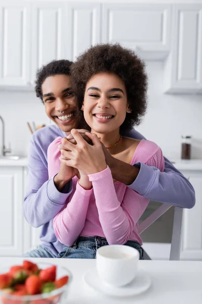 Sonriente afroamericano hombre abrazando novia cerca borrosa taza y fresas en la cocina - foto de stock
