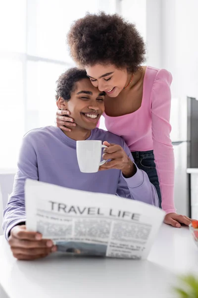 Sorrindo afro-americano mulher abraçando namorado com copo e jornal borrado em casa — Fotografia de Stock