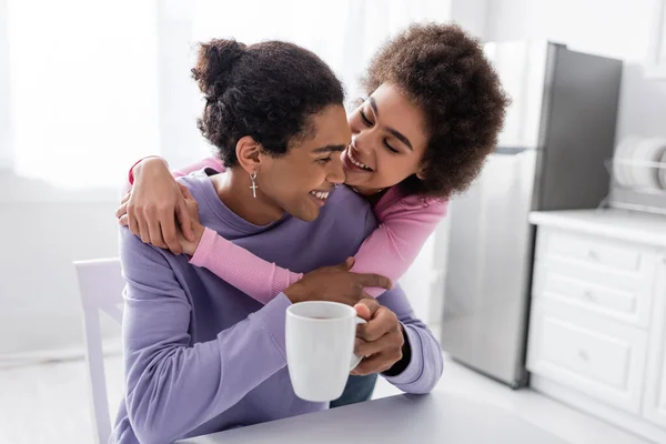 Sonriente afroamericana mujer abrazando novio con taza en cocina - foto de stock