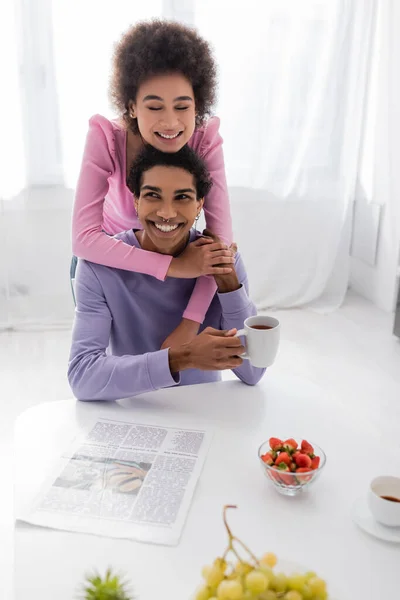Heureuse femme afro-américaine embrassant petit ami avec tasse près de fruits et café à la maison — Photo de stock