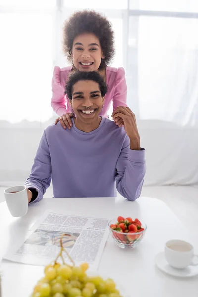 Joven pareja afroamericana sonriendo a la cámara cerca de bebidas, frutas y periódicos en casa - foto de stock
