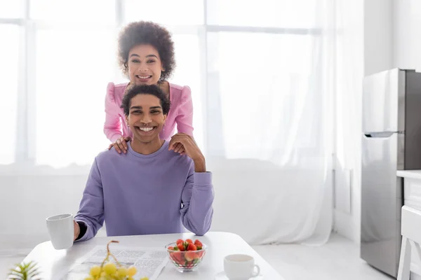 Couple afro-américain positif regardant la caméra près du journal, du café et des fruits dans la cuisine — Photo de stock