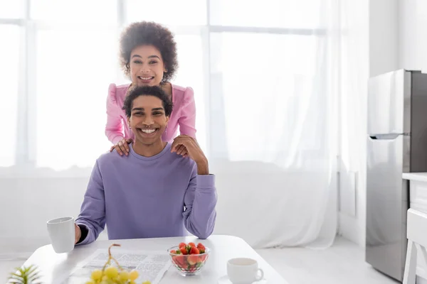 Positive african american couple looking at camera near newspaper, coffee and fruits in kitchen — Stock Photo