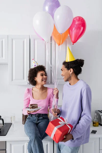 Couple afro-américain souriant avec ballons, cadeau et champagne dans la cuisine — Photo de stock