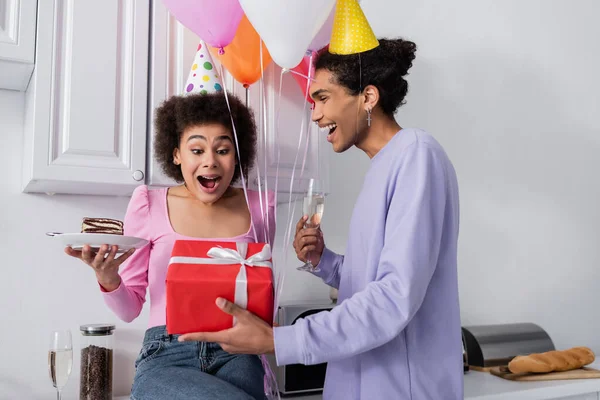 Donna afro-americana eccitata che tiene la torta di compleanno vicino al fidanzato con champagne e regalo in cucina — Foto stock