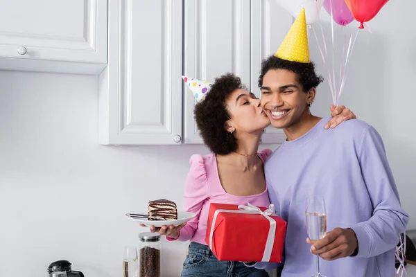 Positive african american woman holding cake and kissing boyfriend with gift and champagne at home — Stock Photo