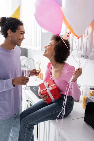 Side view of cheerful african american couple in party caps holding champagne and present at home — Foto stock