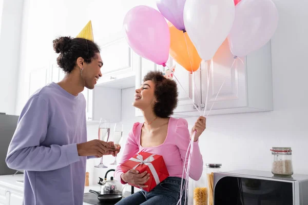 Smiling african american woman holding balloons and gift near boyfriend with champagne in kitchen — Foto stock