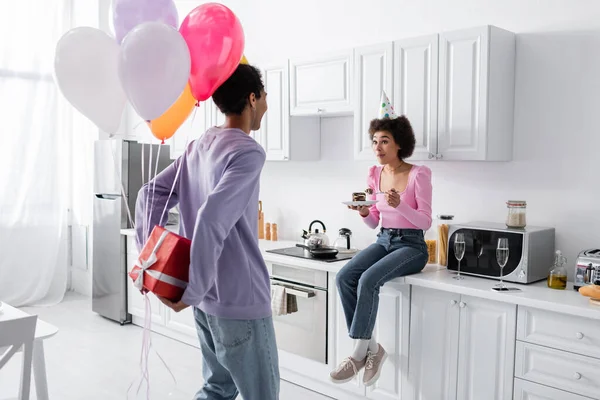 Joven afroamericano hombre escondido presente y globos cerca de novia asombrada con pastel en casa - foto de stock