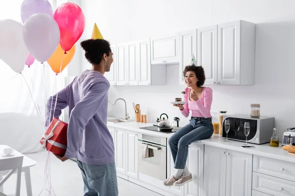 Sonriente mujer afroamericana en gorra de fiesta sosteniendo pastel cerca de novio con regalo y globos en la cocina - foto de stock