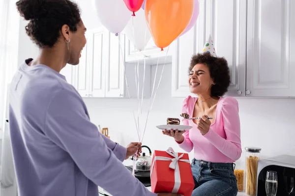 Mujer afroamericana feliz sosteniendo pastel cerca de novio con globos y caja de regalo en la cocina - foto de stock