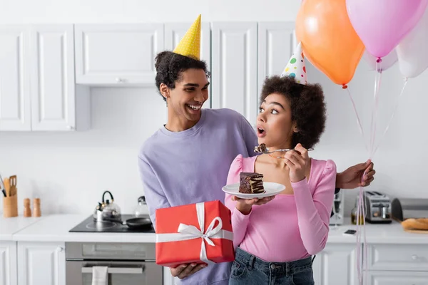 Sorrindo homem afro-americano em boné de festa segurando balões e presente perto da namorada com bolo na cozinha — Fotografia de Stock