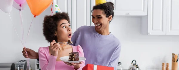 Excited african american woman holding birthday cake near boyfriend with present and balloons at home, banner — Stock Photo