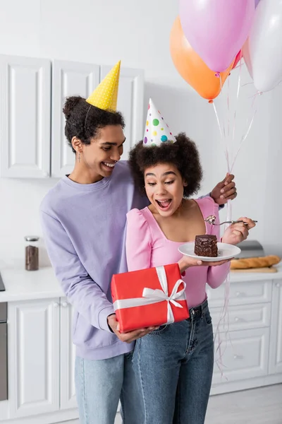 Souriant homme afro-américain tenant présent près de petite amie étonnée dans le chapeau de fête avec gâteau d'anniversaire à la maison — Photo de stock