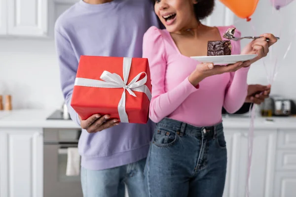 Vista ritagliata del presente in mano dell'uomo afroamericano sfocato vicino alla ragazza con torta a casa — Foto stock