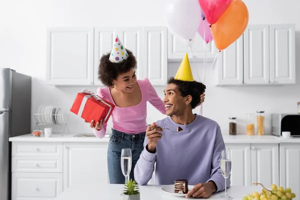 Alegre mujer afroamericana sosteniendo presente cerca de novio en la tapa del partido y pastel de cumpleaños en casa - foto de stock